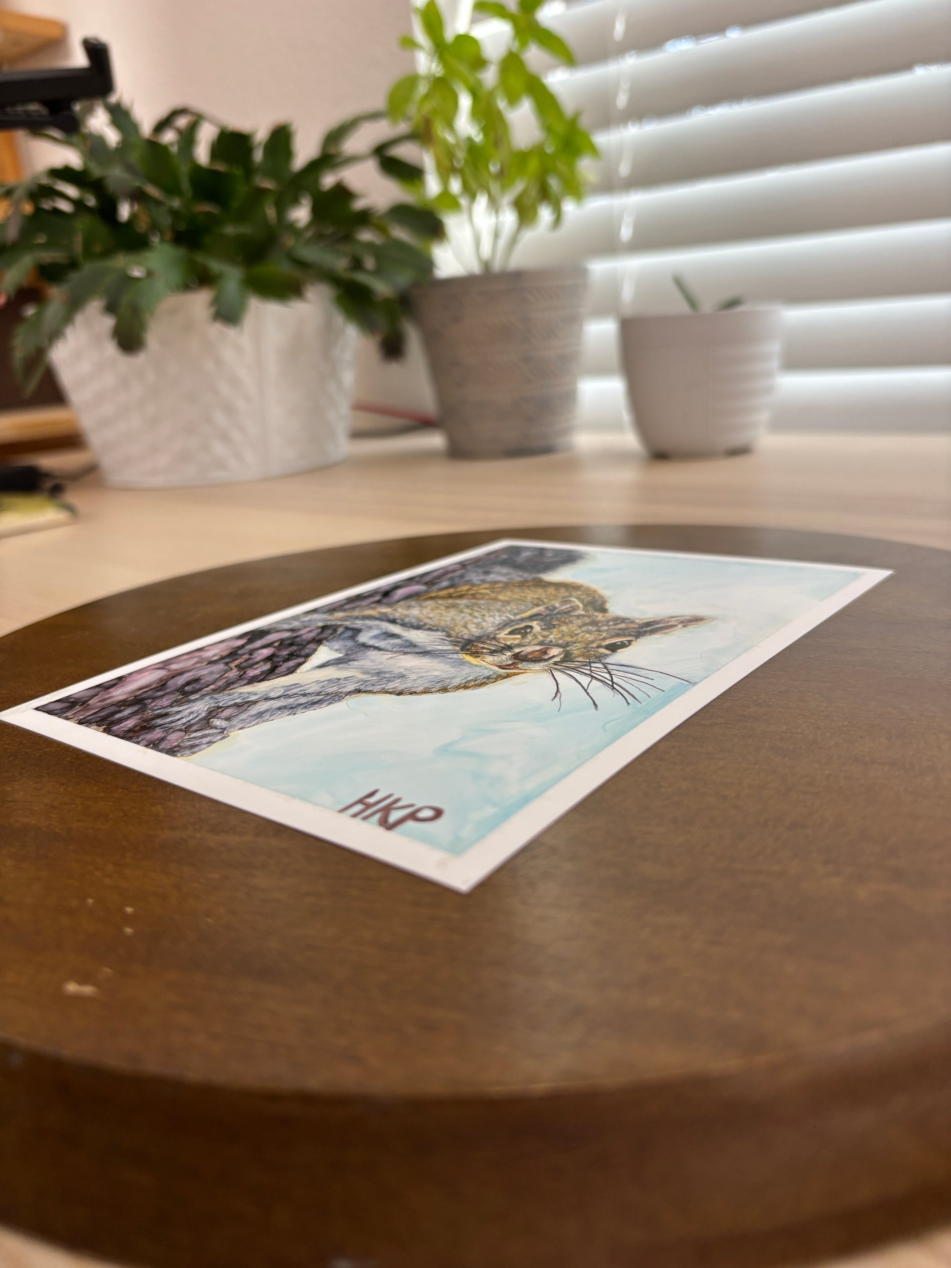 Painting of a squirrel from the side, showing that it is painted on flat paper. Some plants are shown in the blurred background.